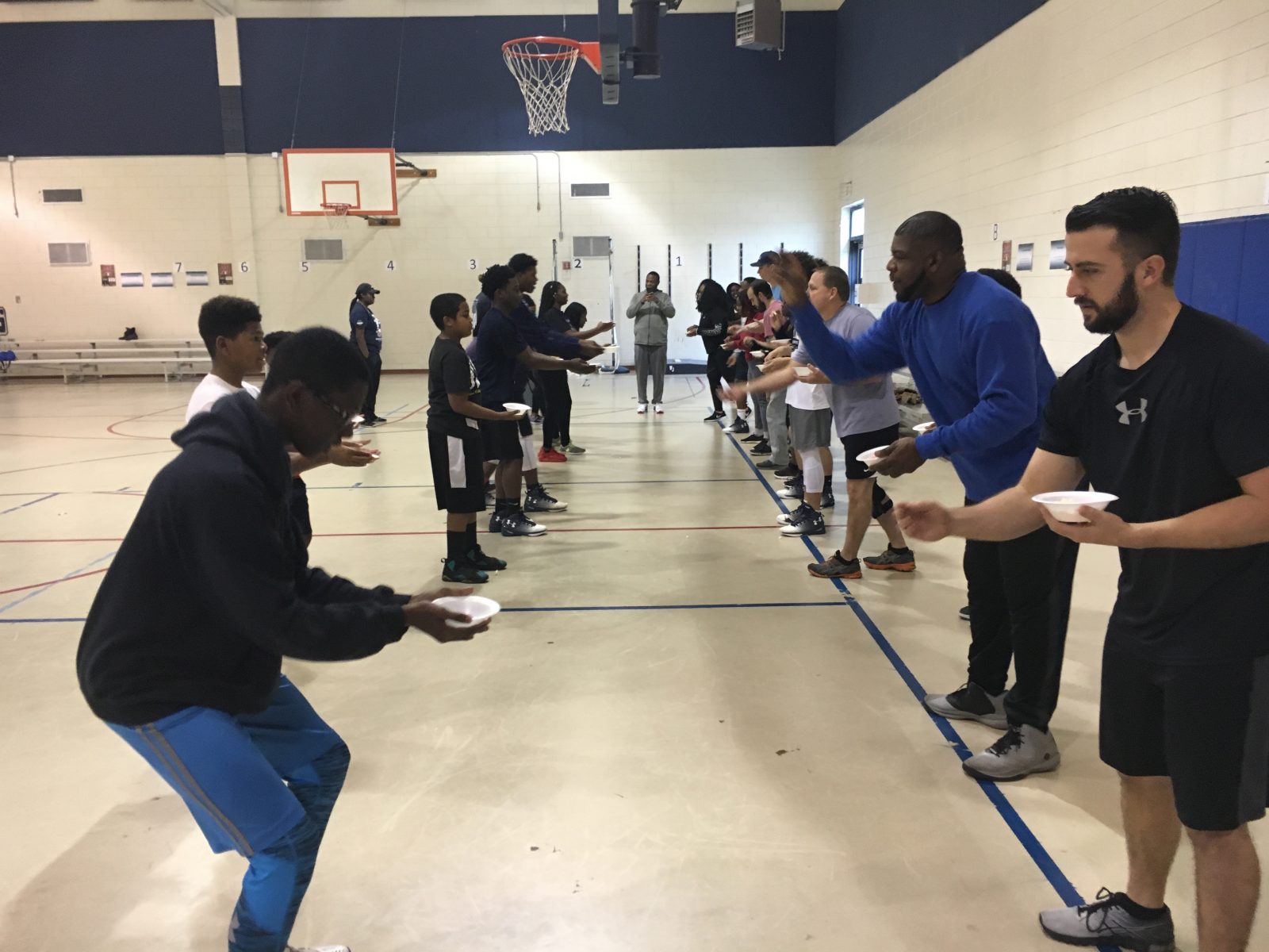 Group of children and adults standing in two lines facing each other on an indoor gymnasium floor participating in a team-building activity or recreation game.