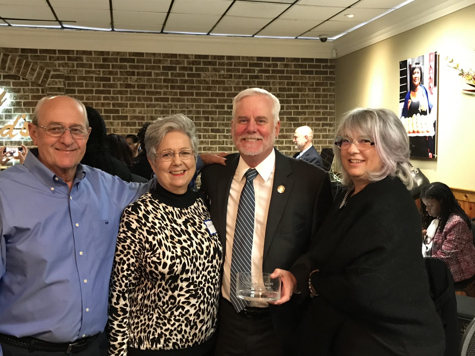 A group photo of four smiling adults at an indoor reception or event, with one holding a glass award.