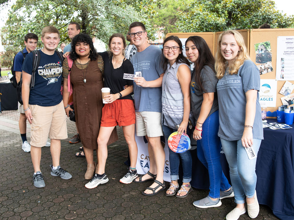 A group of seven smiling young adults pose together outdoors at an event, some holding flyers and wearing casual clothes. Display boards with information and event materials are visible in the background.