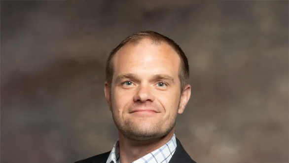 Professional headshot: smiling man in a blazer and light checkered shirt against a neutral studio backdrop.