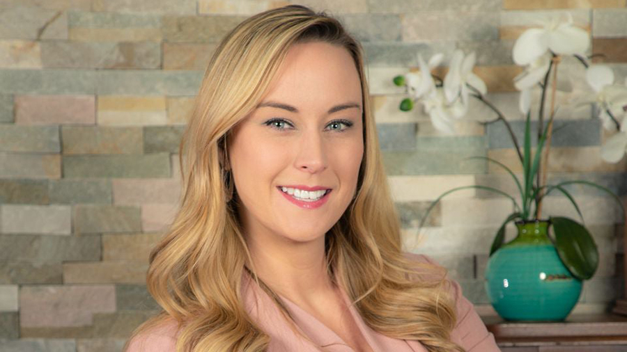 Portrait of a smiling blonde woman in a pale pink top, with a stone-tile wall and a turquoise vase with white orchids behind her.