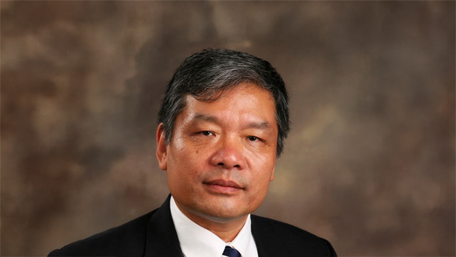 Portrait of a middle-aged man in a dark suit and white shirt against a brown studio background with a neutral expression.