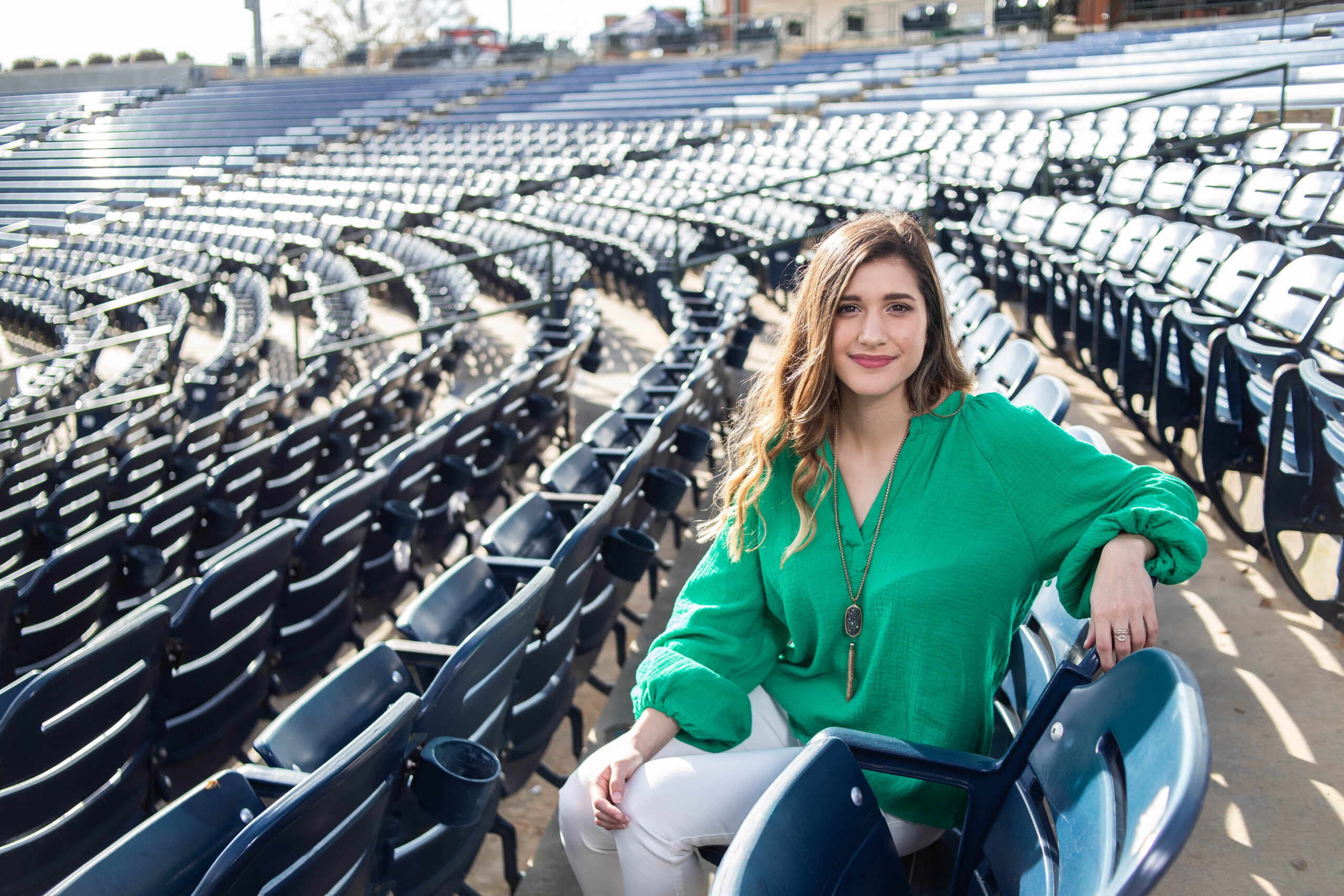 A smiling young woman wearing a bright green blouse and long pendant necklace sitting in empty stadium seating.