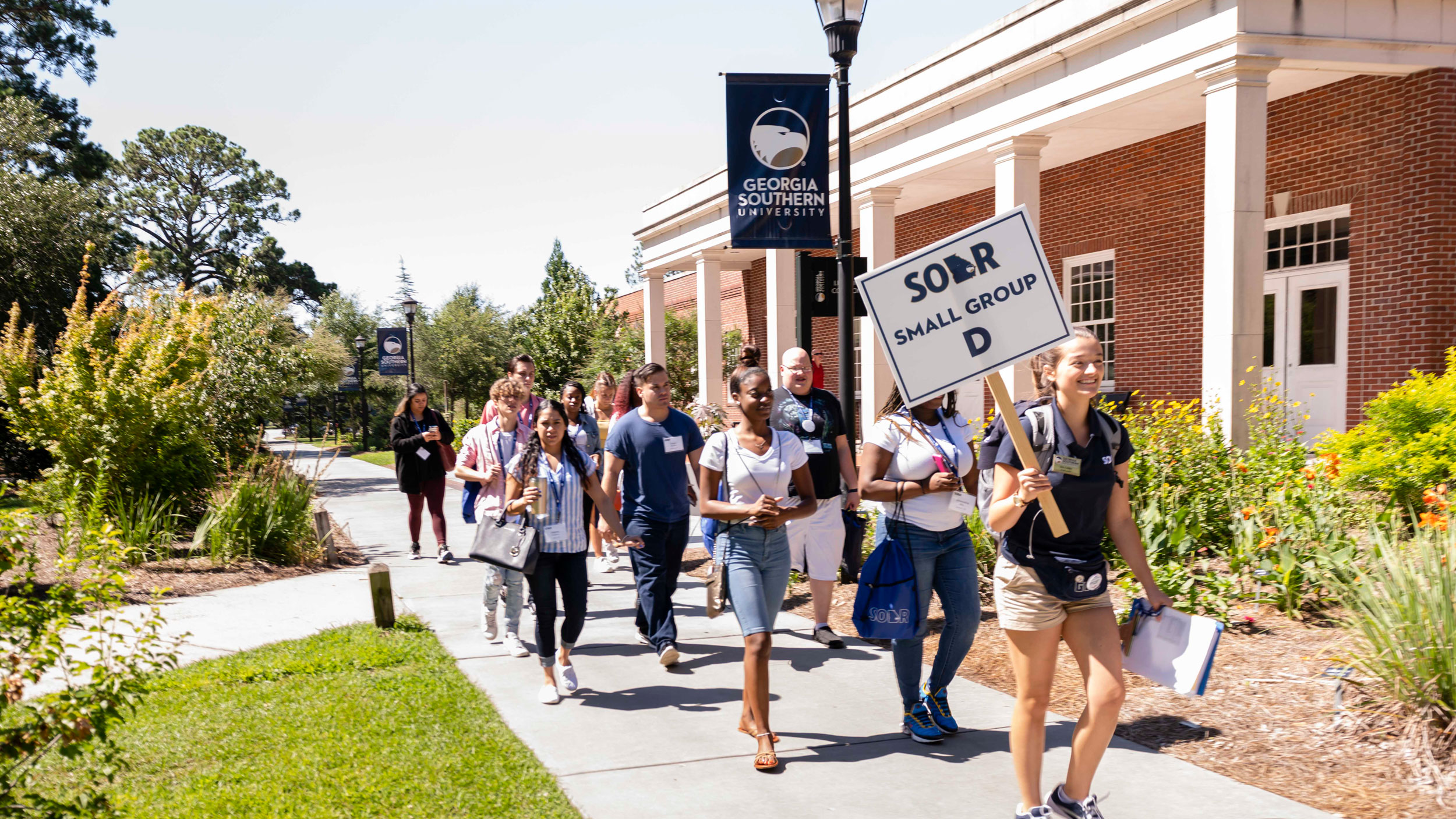 A group of students walks outside on a campus sidewalk, led by a person holding a SOAR Small Group D sign at Georgia Southern University. They are surrounded by greenery and a brick building.