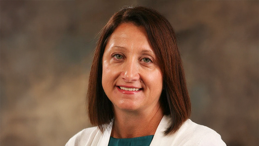 Portrait of a smiling woman with shoulder-length brown hair wearing a white blazer over a teal blouse, against a neutral studio backdrop.