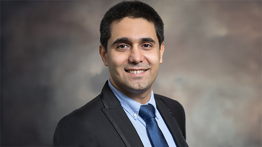 Smiling man in a dark plaid suit, blue shirt, blue tie, posed against a brown studio backdrop (professional headshot)