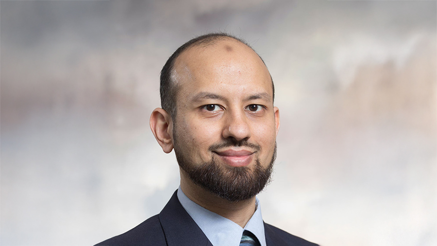 Professional headshot of a man with a trimmed beard wearing a navy suit and light blue shirt, smiling softly.