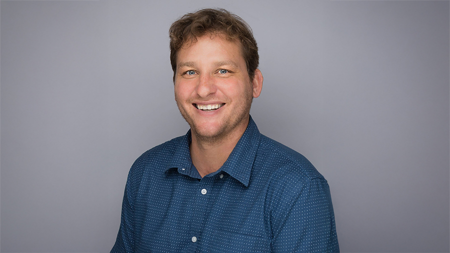 Smiling man with short brown hair in a blue button-down shirt against a gray backdrop.