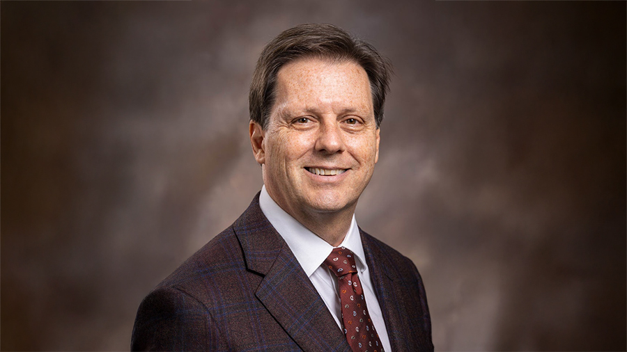 Smiling man in a dark plaid suit, white shirt, red tie, posed against a brown studio backdrop (professional headshot)