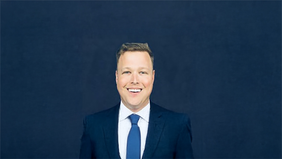 Professional headshot: smiling man in a navy suit and blue tie against a dark blue backdrop, suitable for business profiles.