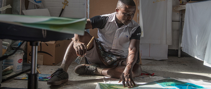 Male artist sitting on the studio floor holding a paintbrush and working on a painting canvas.