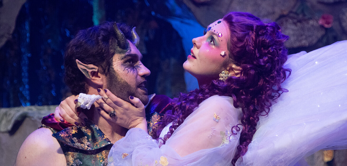 Two theatrical actors in mythical costumes, one as a horned creature and the other with purple hair and fairy wings, performing on stage.