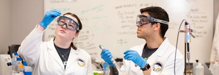 Two researchers wearing lab coats, safety goggles, and blue gloves conducting an experiment in a science laboratory.