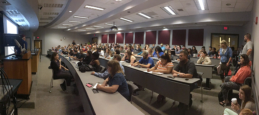 Large group of college students sitting at curved desks in a lecture hall listening to a university presentation.