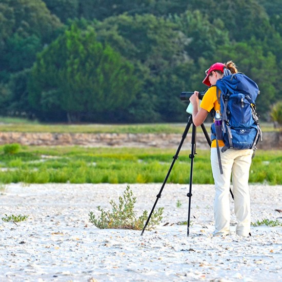 Hiker with a blue backpack using a spotting scope on a tripod to observe wildlife or a landscape near a green field.