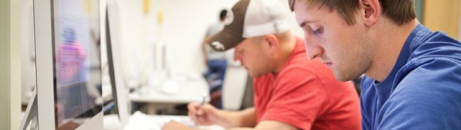 Two male students working intently at computer monitors in a university computer lab or classroom setting.