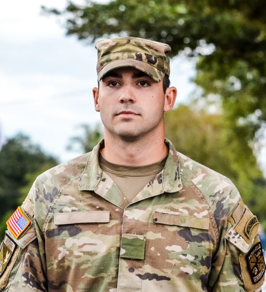 Close-up portrait of a US Army service member in a camouflage uniform and cap with outdoor trees in the background.