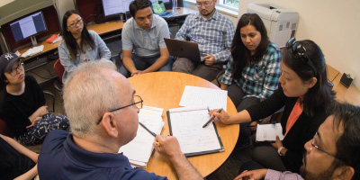 A group of university students gathered around a circular wooden table listening to an older professor point at notes on a clipboard.