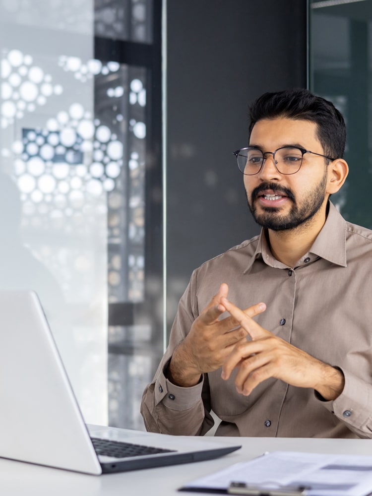 A man with glasses and a beard sits at a desk, gesturing with his hands while looking at a laptop. Papers and a pen are on the desk, and a window with geometric patterns is in the background.
