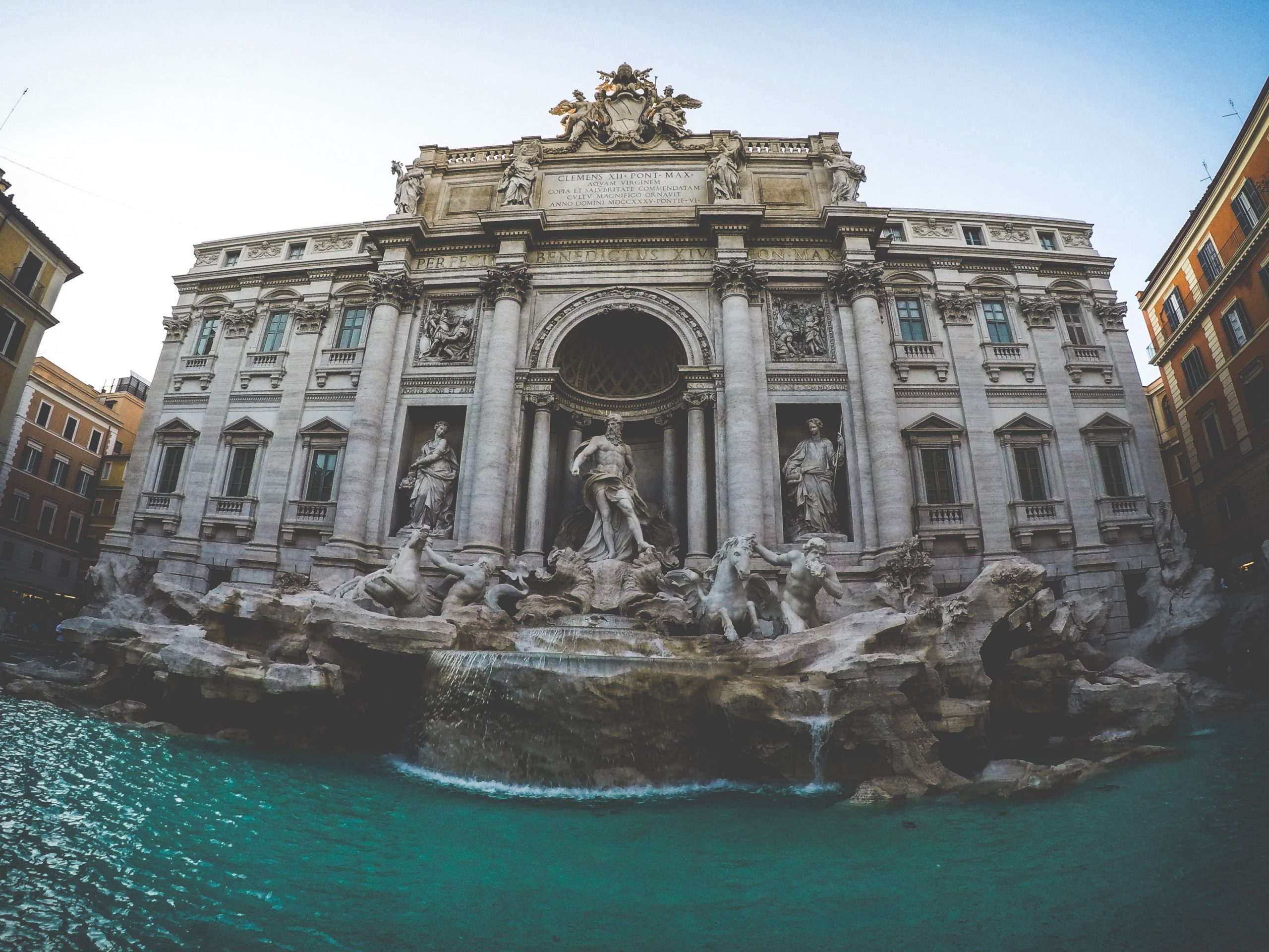 The Trevi Fountain in Rome, Italy, featuring an ornate Baroque façade with large marble statues, cascading water, and a turquoise pool in the foreground.
