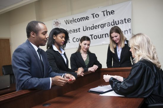 Five students dressed in business attire participate in a mock courtroom competition, standing before a judge at a courtroom bench beneath a tournament welcome banner.