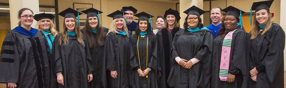 Diverse group of university graduates and faculty posing in black academic regalia and caps at a graduation ceremony.