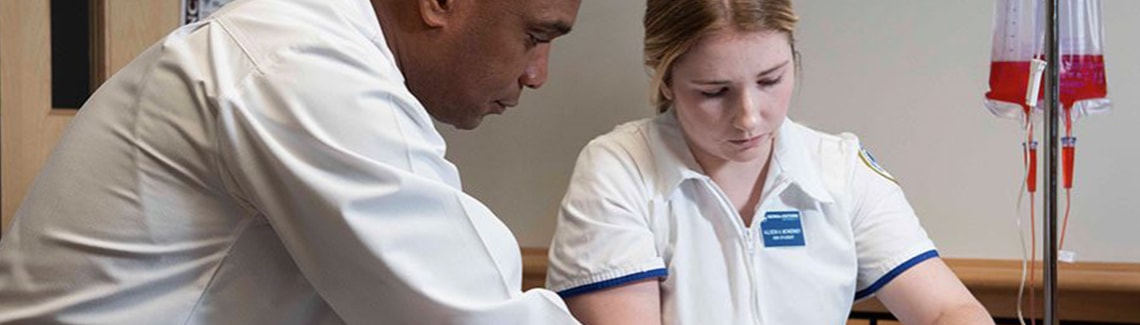 A male medical instructor guiding a female nursing student during a training session next to an IV stand.