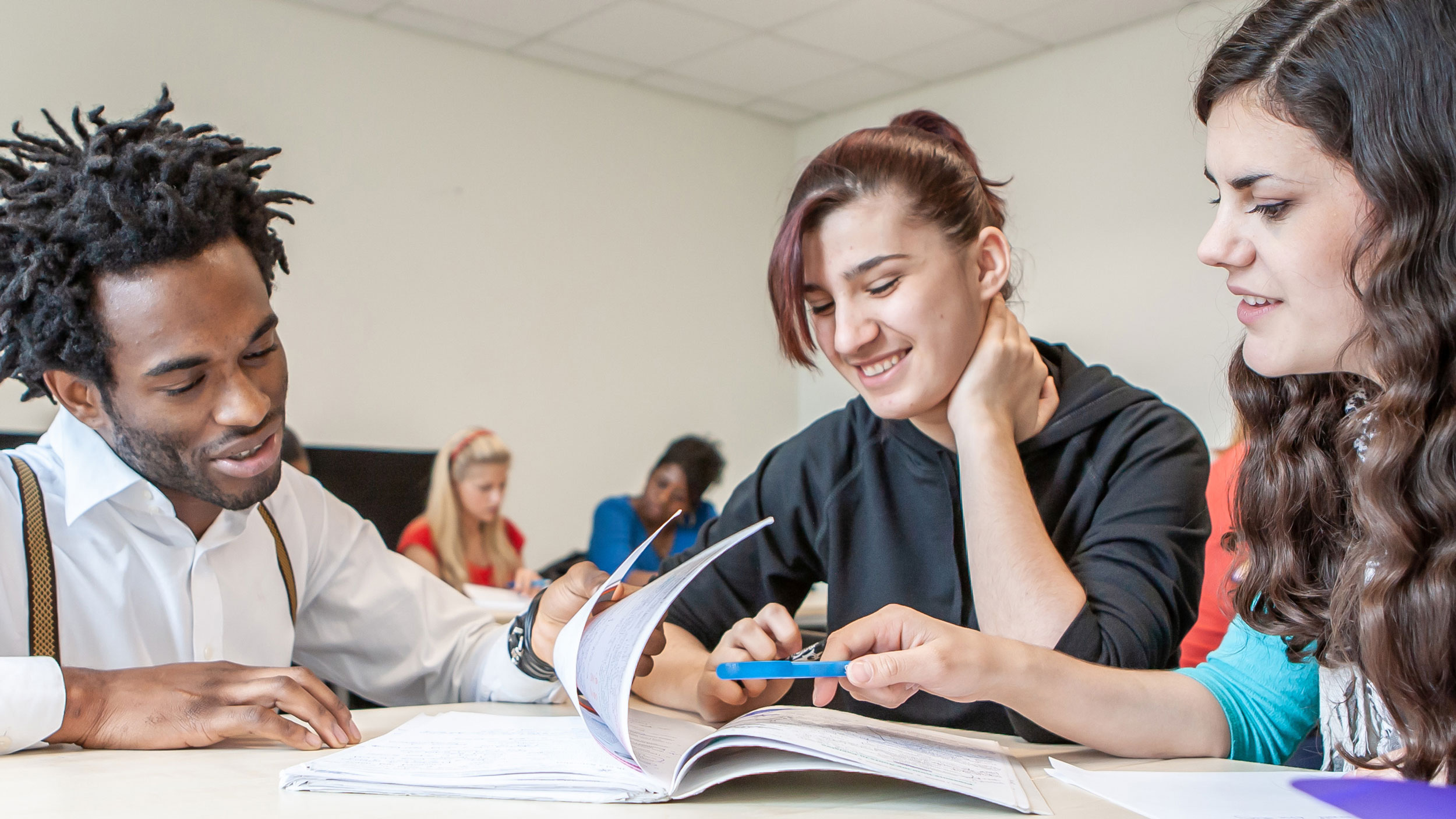 A male instructor with dreadlocks wearing suspenders and a white shirt working with two female students who are reviewing printed study materials at a desk in a classroom.
