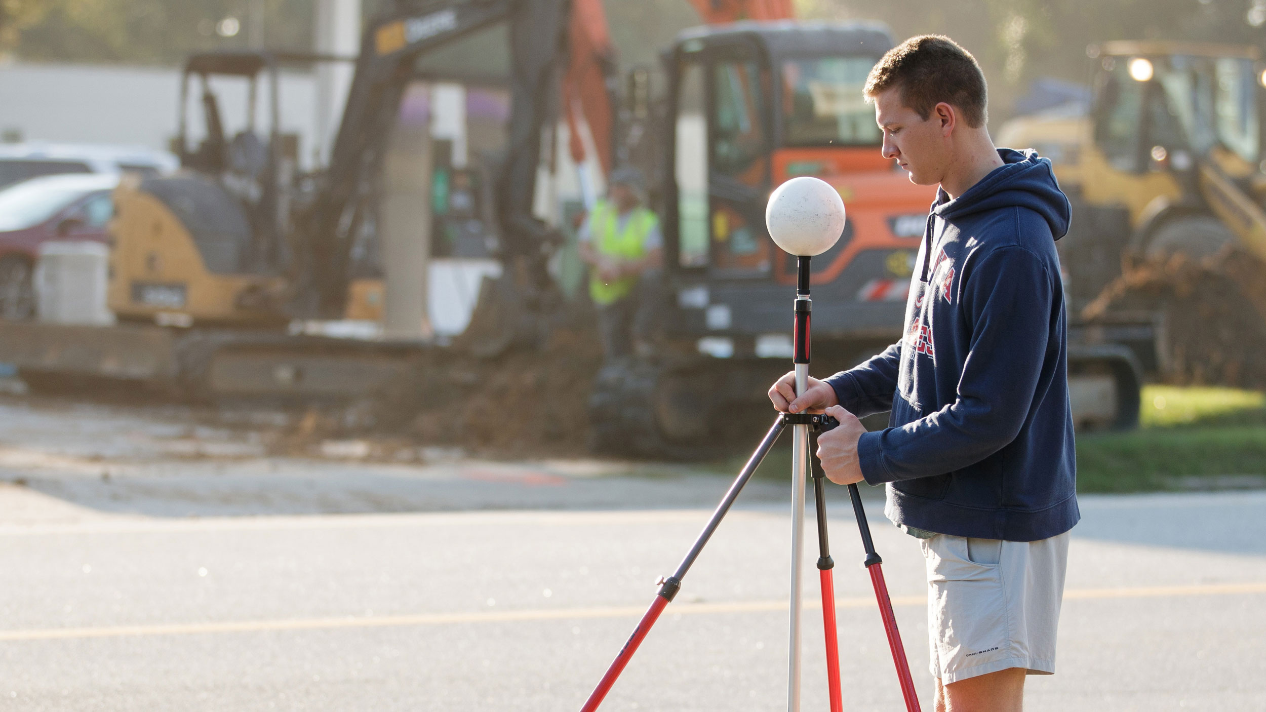 A male student operating land surveying/GPS sphere target equipment on a tripod near a construction site with heavy machinery.