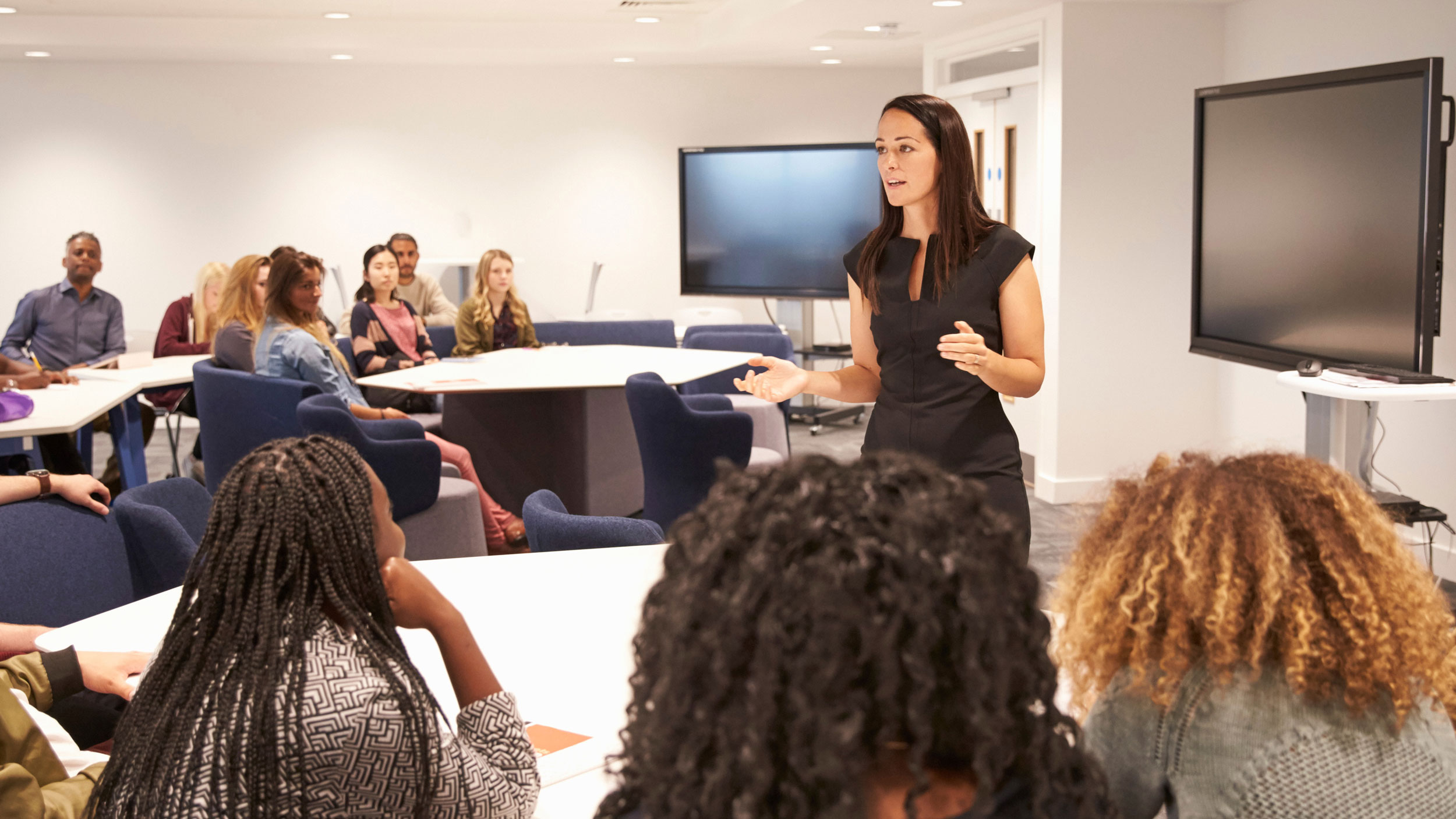 A female instructor in a black dress speaking gesturingly to a group of students seated around white tables in a modern classroom setting.