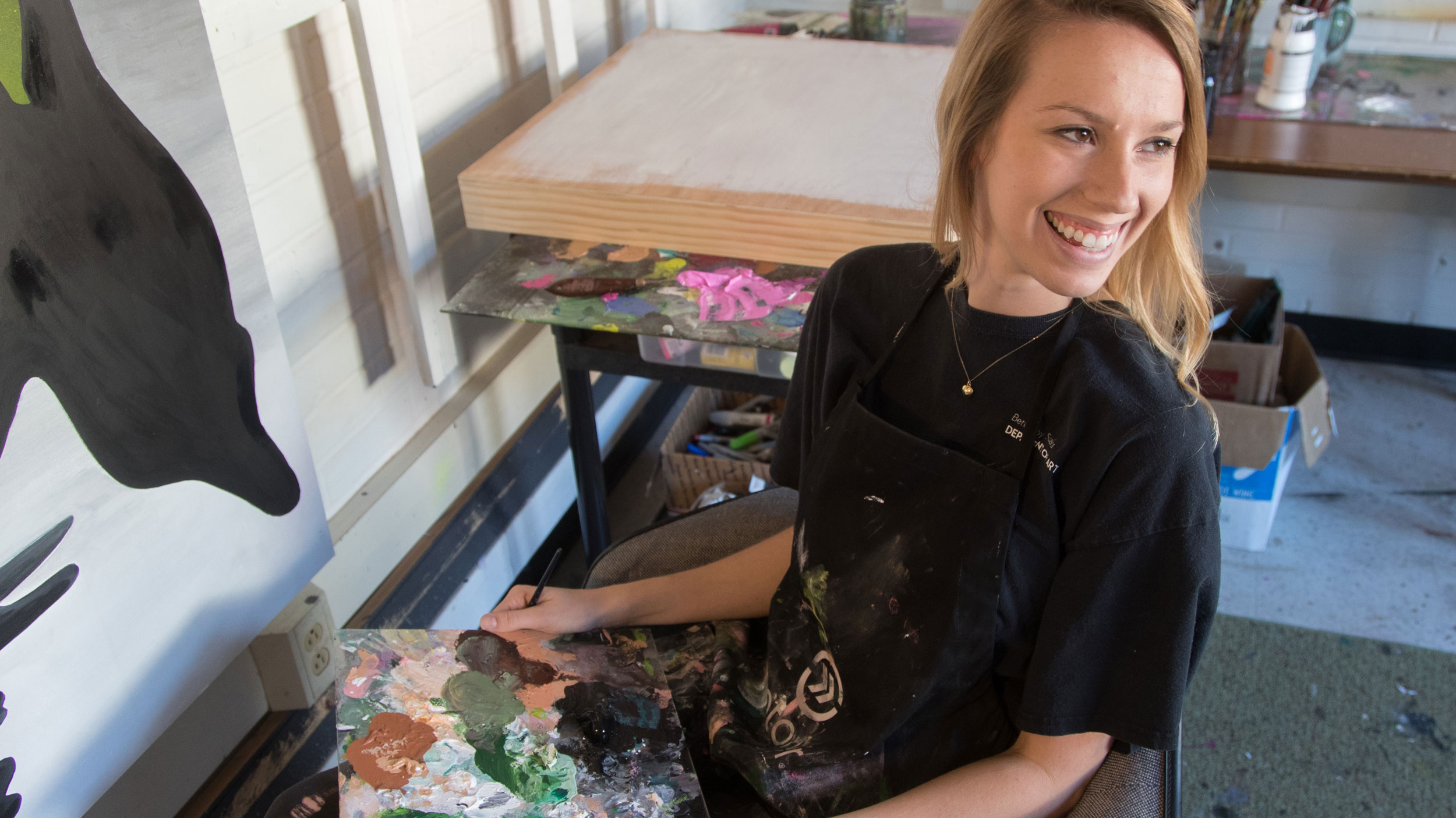 A smiling female student wearing a paint-splattered black apron, holding a paint palette and brush while sitting in an art studio.