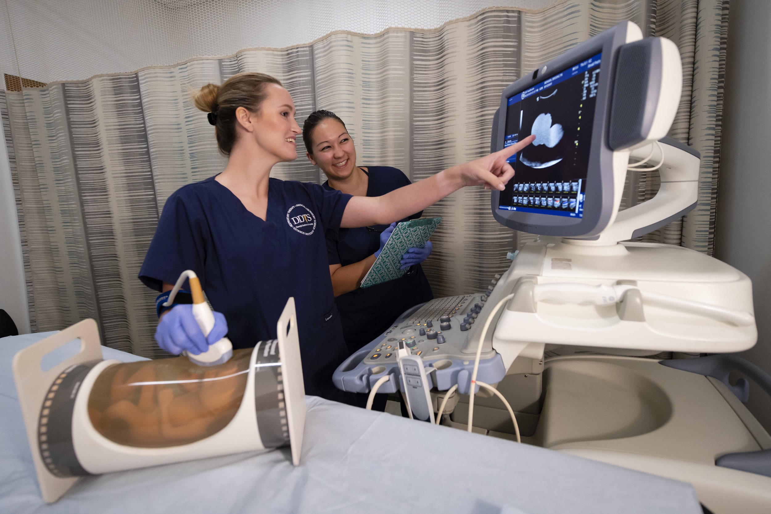Two women in medical scrubs use an ultrasound machine, with one pointing to the monitor and the other holding a notepad. A medical training mannequin is on the table beside them.