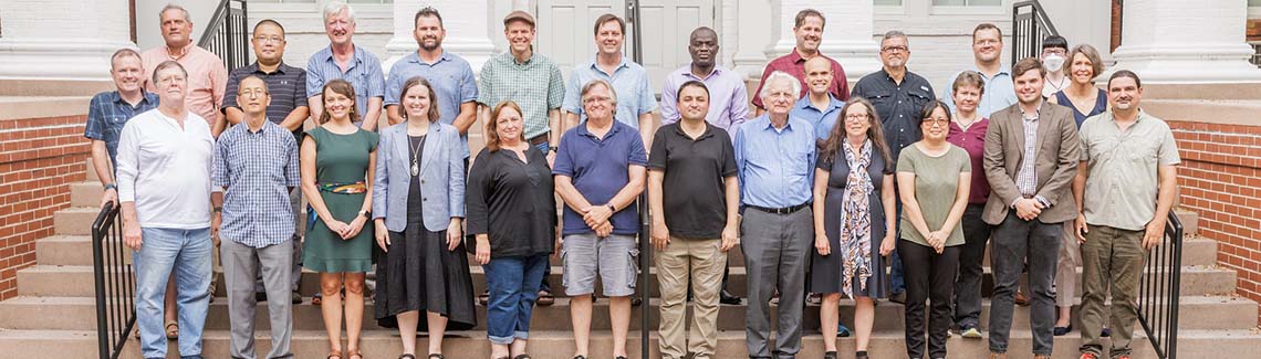 A large group photograph of university faculty members and staff standing together on the outdoor brick steps of a classic campus building with white pillars.