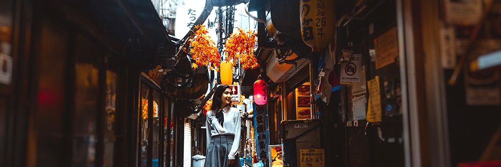 A narrow, atmospheric Japanese alleyway decorated with vibrant orange autumn foliage overhead, paper lanterns, traditional storefront signs, and a smiling woman walking in the center.