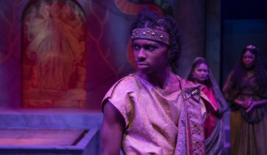 A male actor with dreadlocks wearing a patterned gold tunic and beaded headband, looking back over his shoulder in a theatrical performance. Two other actors in tunics stand in the dimly lit, purple-hued background.