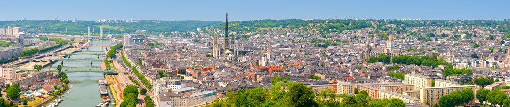 A panoramic skyline view of Rouen, France, showing the Seine River winding past bridges with the spire of the Rouen Cathedral rising in the center.
