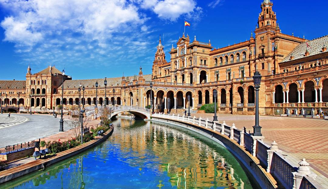 A wide, vibrant shot of the historic Plaza de España in Seville, Spain, featuring its iconic semicircular pavilion building, arched bridges, and canal