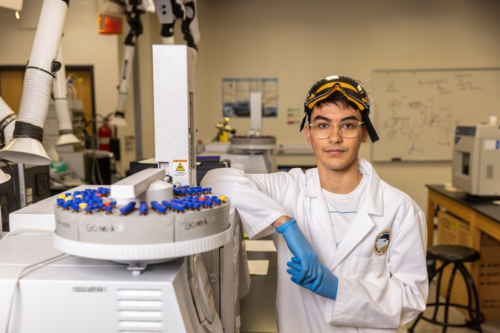 A portrait of a male student researcher wearing a white lab coat with a Georgia Southern logo patch, safety goggles resting on his head, blue nitrile gloves, and clear glasses,