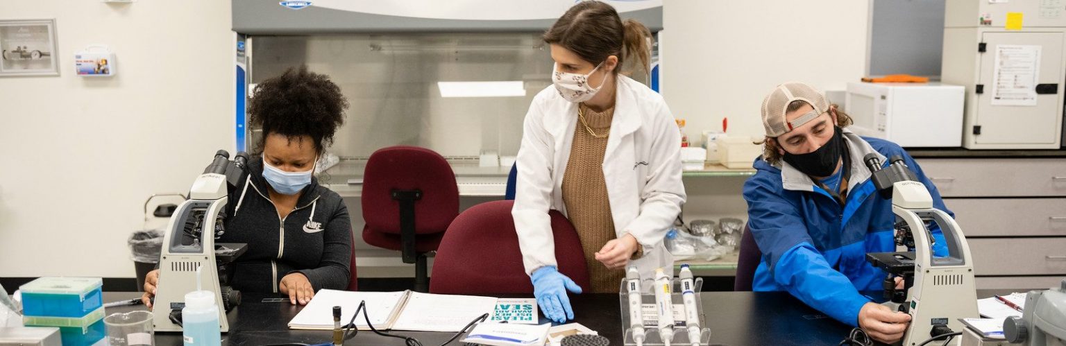 A laboratory scene showing a masked female lab instructor standing between two students wearing face masks—one student on the left leaning over a microscope, and a male student on the right adjusting his microscope, with lab equipment and pipettes on the desk.