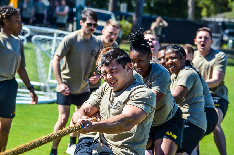 An energetic outdoor photo of Army ROTC cadets in matching olive athletic shirts intensely pulling on a thick rope during a tug-of-war competition on a sunny lawn.