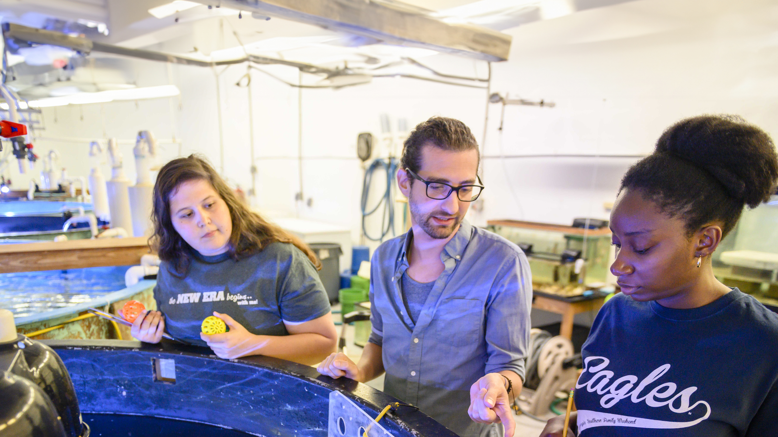 A science laboratory shot showing a male instructor or researcher with glasses speaking to two female students gathered around a large aquatic tank. One student holds colorful plastic enrichment objects while the other listens closely in a navy 