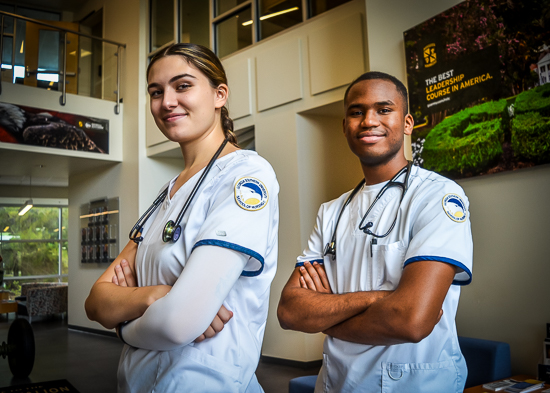 A portrait of two Georgia Southern nursing students—a female and a male—standing side by side with crossed arms, wearing white nursing scrubs with school patches and stethoscopes around their necks inside a campus building.