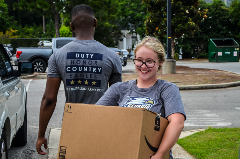 A smiling female student carrying a large cardboard box on move-in day, with a male student walking beside her wearing a gray t-shirt that reads 