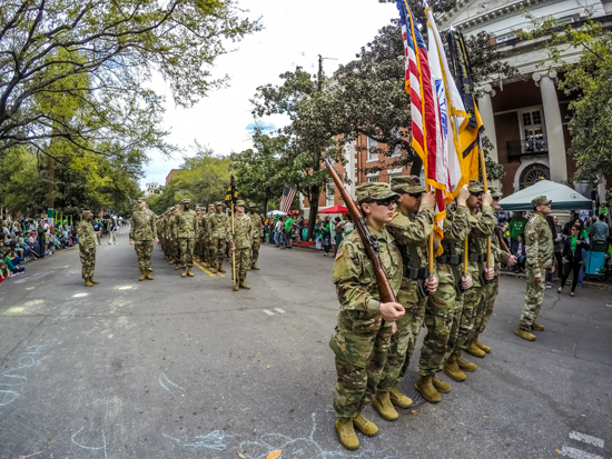 An Army ROTC color guard and formation marching in a street parade lined with spectators in front of historic brick buildings.