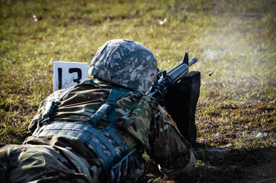 An Army ROTC cadet lying in a prone position on a outdoor shooting range, aiming a rifle during a live-fire weapons qualification or marksmanship training, with a spent casing flying through the air.