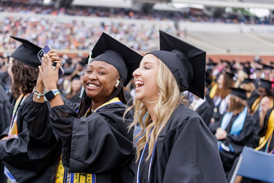 Two smiling female graduates in black caps and gowns taking a selfie with a smartphone during an outdoor stadium graduation ceremony.