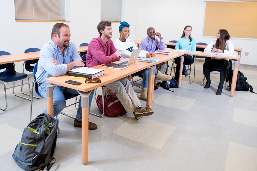 A group of diverse adult students seated at desks arranged in a semi-circle during an engaging classroom discussion.