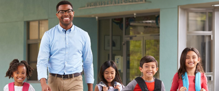 A smiling male teacher with glasses in a light blue button-down shirt walking outdoors in front of a school building with four young elementary students wearing backpacks.