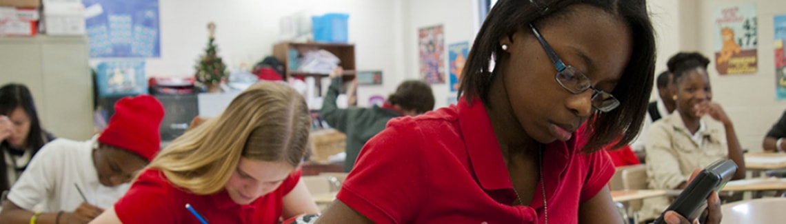 A close-up view of a female student with glasses in a red polo shirt using a handheld electronic device at her desk during a busy classroom session.