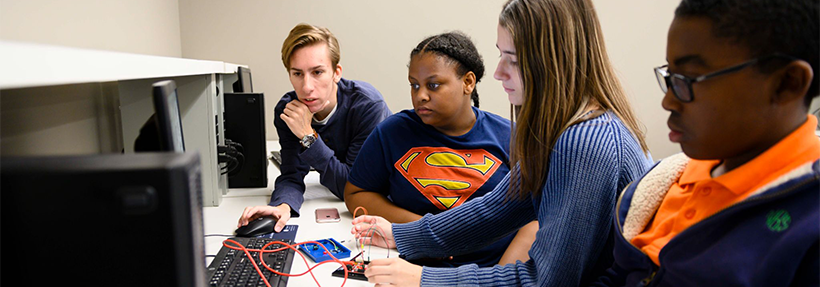 A group of four students collaborating in a computer lab setting; one male student leans over a mouse and keyboard while others focus on testing electronics, breadboards, and wiring at a desk station.